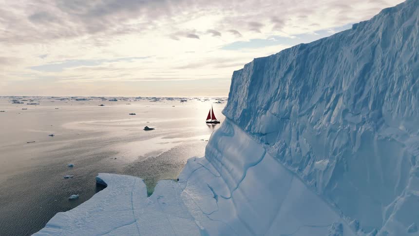 A broad sheet of broken sea ice glows pale blue under soft clouds as a sailboat floats along