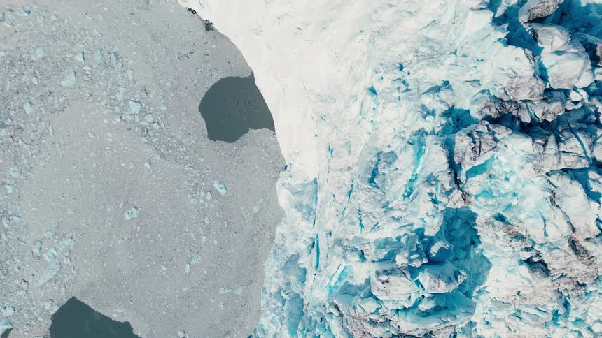 The video is a highangle drone shot looking straight down on a massive bright blue and white glacier the glaciers heavily crevassed icy surface contrasts sharply with the dark gray water and loose fragmented ice floating alongside it