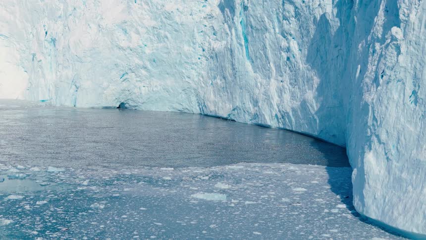 A towering blue glacier wall curves around a quiet cove of dark water