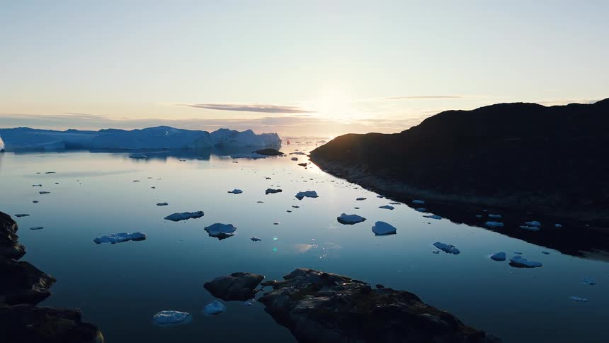 An aerial wide shot shows a woman in a red jacket walking on a rocky mountain path overlooking a dark fjord filled with floating icebergs as the sun sets over the horizon