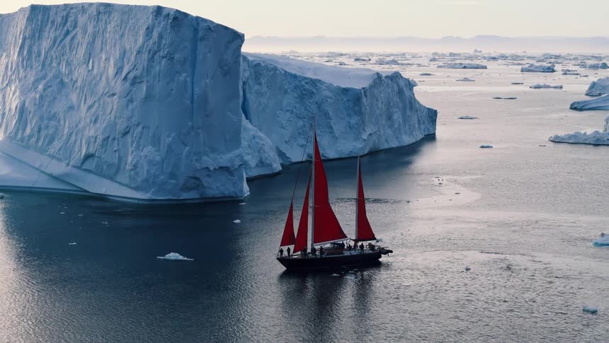 A twomasted sailboat with red sails navigating through a body of water dotted with ice floes passing a vast sheer wall of bluewhite ice with a field of icebergs stretching toward the horizon