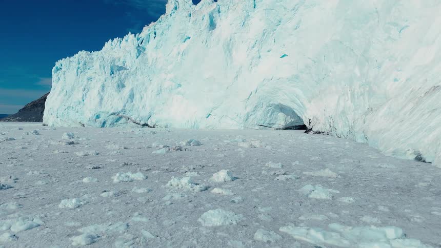 A vast field of fractured sea ice stretches toward a towering bright white glacier wall with a dark cave like opening under a clear deep blue sky