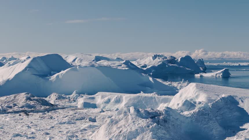 The video provides an expansive aerial view of a rugged snowy landscape featuring a large accumulation of icebergs the scene is dominated by massive sculpted formations of white and blue ice under a bright blue sky