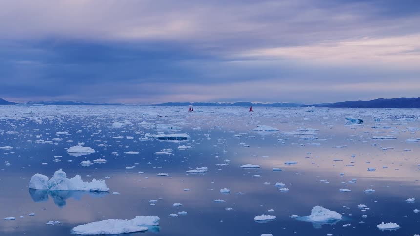 Small red sailboat sails through icy arctic waters under a blue gray sky
