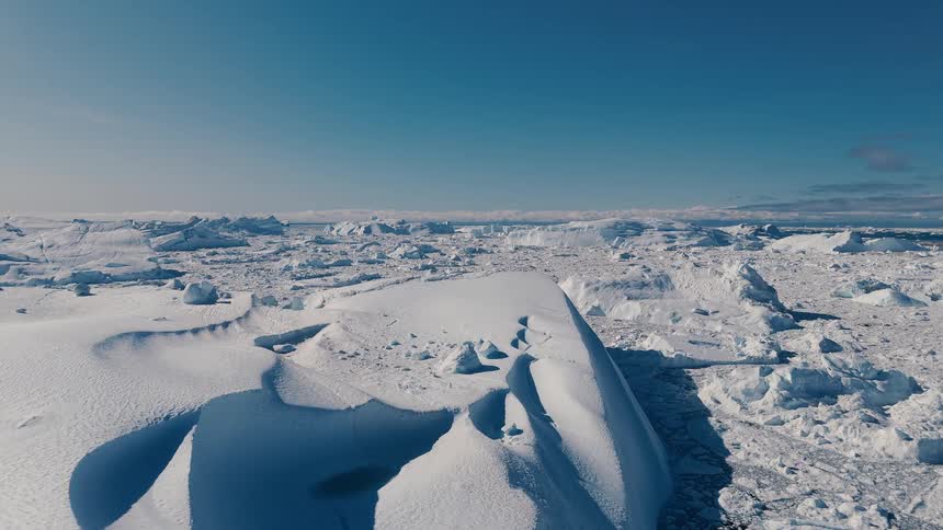 A camera moves over a large expanse of white ice and snow passes over the fractured top of a tall bluewhite glacier wall and reveals a field of smaller ice fragments extending toward a clear blue sky