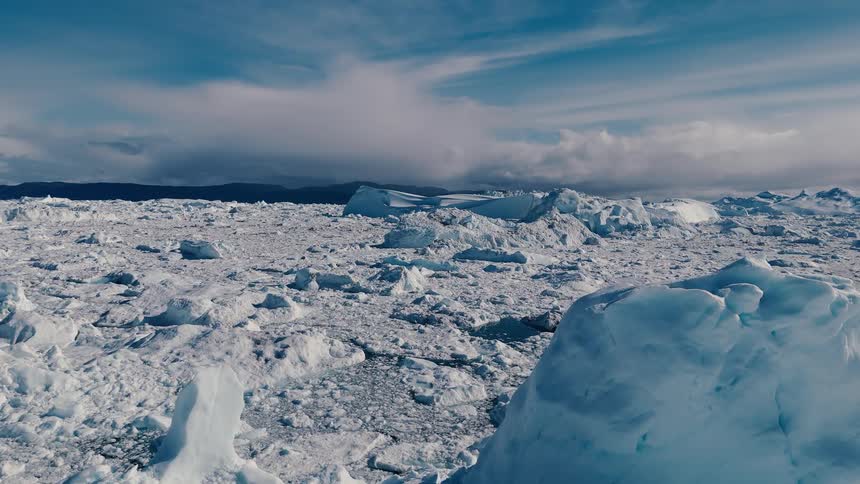 An aerial camera shot moves forward over a densely packed field of broken icebergs and glacial debris with a dark blue and white cloudy sky visible above the wide expanse of ice