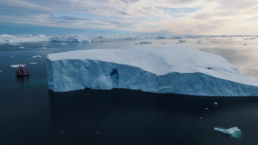 A massive flat topped iceberg floats on calm dark water beneath layered clouds with a sailboat peeking along its left edge