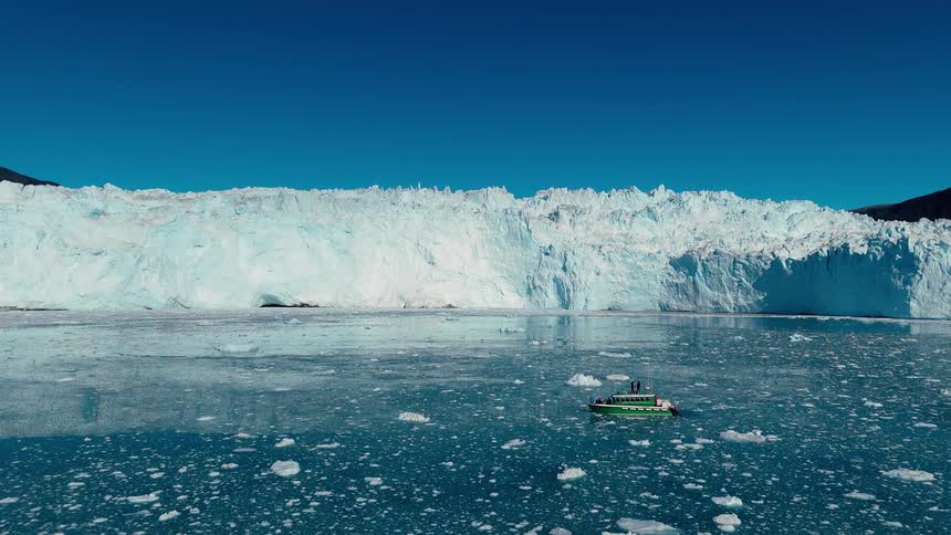The video is an extreme long shot showing a massive white glacier wall meeting the dark water with a small boat visible in the middle distance the bright blue sky and sunlit glacial ice contrast with the dark rock of the surrounding mountain