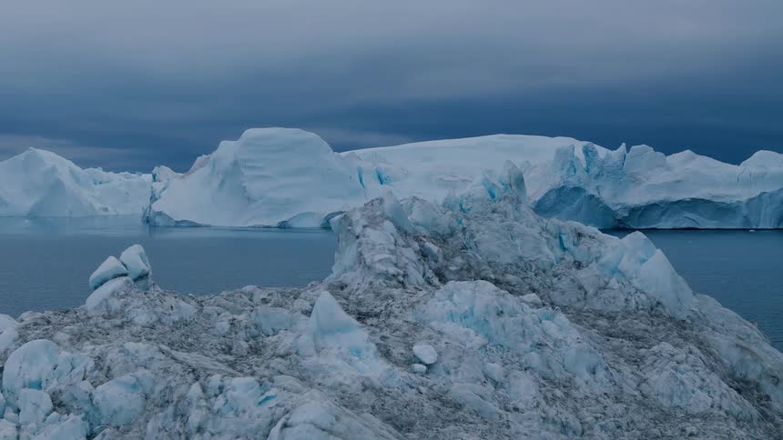 An aerial view of the cold ocean shows a dark textured iceberg in the foreground slowly descending to reveal a massive smooth wall of glacier ice in the distance under a dark gray sky