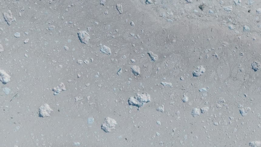 An overhead camera looks down upon a field of varioussized ice fragments and small icebergs floating on a dark water surface slowly panning across the scattered frozen material