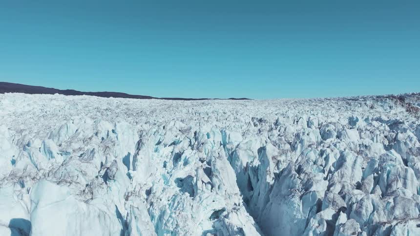 A dramatic aerial view shows the immense fractured blue and white surface of a glacier filling the foreground under a clear bright blue sky with dark mountain peaks in the distance