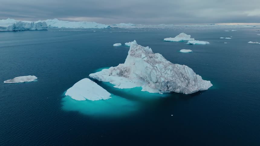 An aerial view of the dark blue ocean shows a massive jagged iceberg with a glowing turquoise base surrounded by other ice fragments under a dramatic cloudy sky