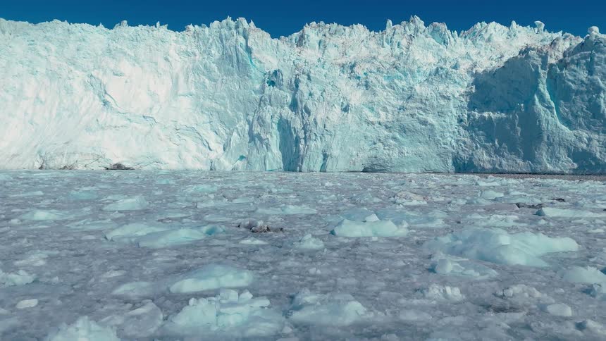 A colossal vertical mass of blue and white ice dominates the background a flat expanse of broken ice pieces covers the water surface and a clear blue atmosphere extends above the top edge of the ice formation