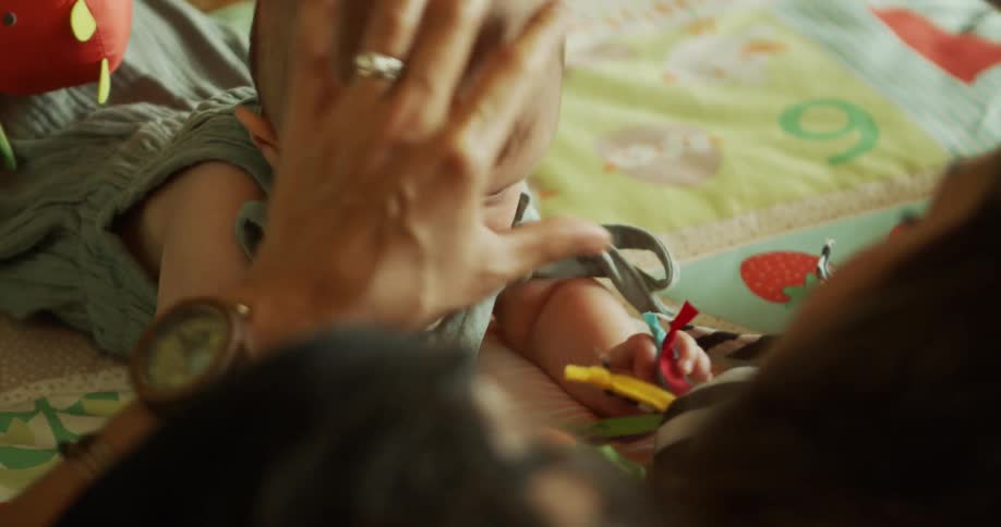 An infant lies on a patterned activity mat the childs clothing has knotted shoulder straps and her mother is gently touching her head in an indoor setting