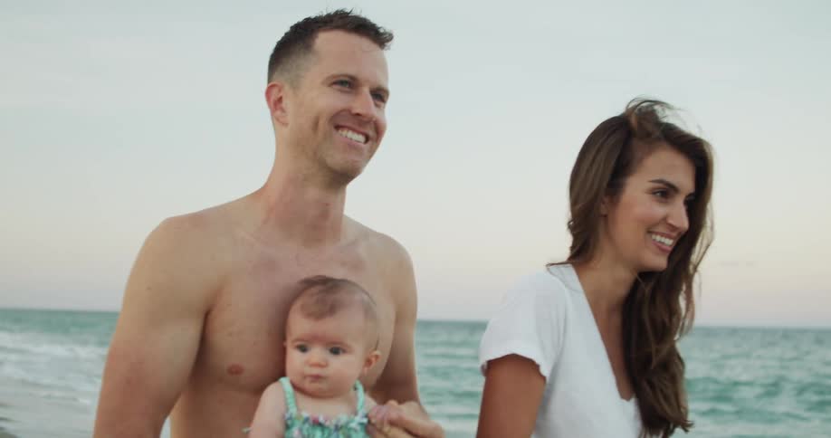 A smiling couple walks along a beach with their infant baby at dusk