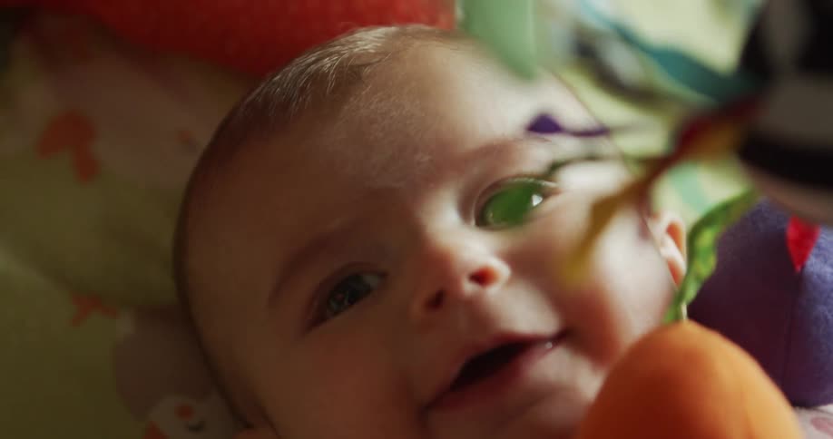 The video is a closeup shot of a babys face lying down with their eyes looking up towards something above themcolorful mobile toys are dangling and slightly out of focus in the foreground indicating the source of the babys attention