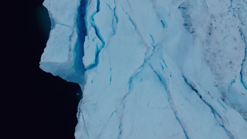 A close high view highlights the sheer fractured blue and white face of a massive glacier or iceberg against a pitch black background