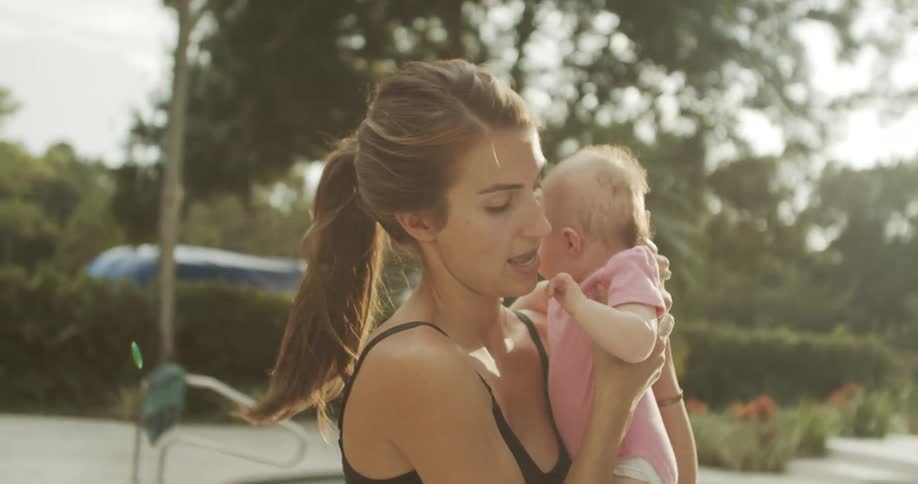 A woman wearing a dark tank top lifts an infant out of a stroller while standing next to a pool structure holds the child close to her chest and looks down at the baby in a sunny yard with trees