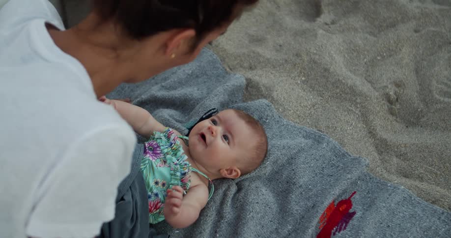 Mother leans over laughing baby lying on the beach sand