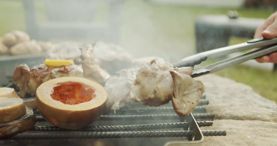 A hand uses tongs to turn sizzling meat beside halved squash on a smoky outdoor grill