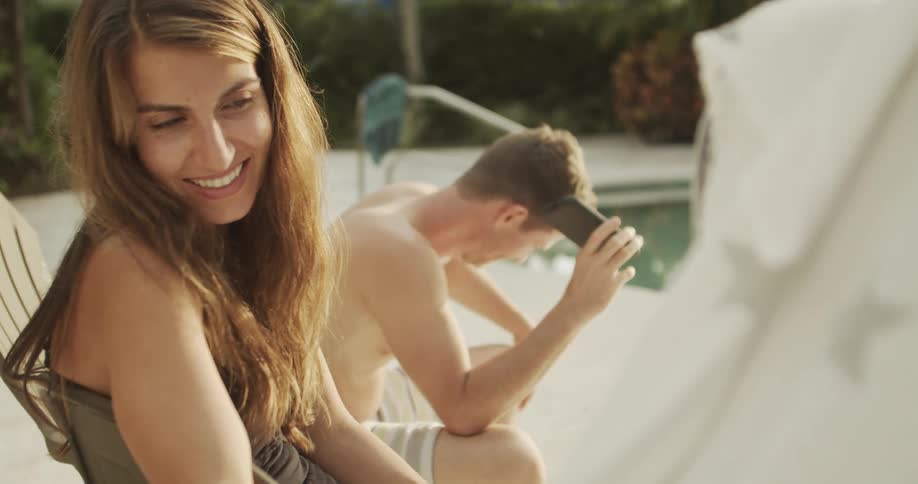 A woman with long brown hair smiles brightly as she sits in a chair in the foreground enjoying a sunny day outdoors in the blurred background a man shirtless and wearing swim trunks is sitting nearby and looking down at his mobile phone