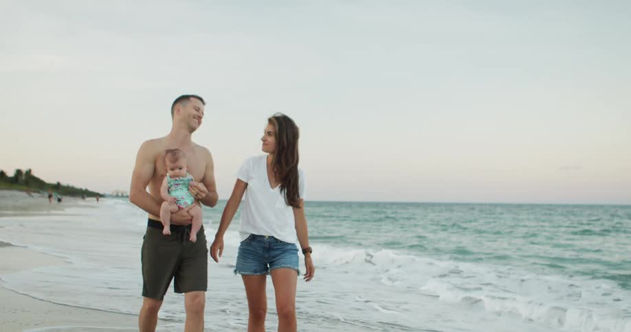 A family walks on a sandy shore the man holds a baby and the woman walks next to them and ocean waves crash near the feet of them under a light atmosphere