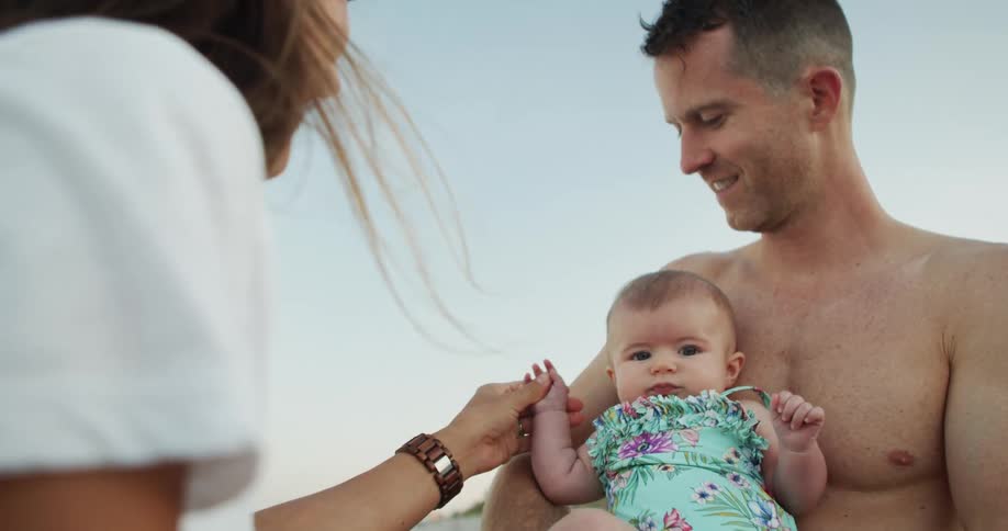 A man holds an infant in a swimsuit a woman leans in toward the child and man the woman holds the hand of the child the man smiles at the people and all are positioned against a bright sky in a beach