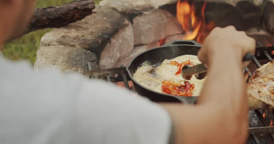 Person cooking meat and peppers in a cast iron skillet over fire
