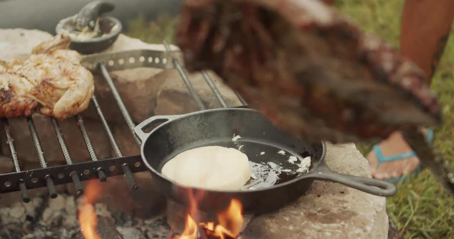The video shows a closeup of a person cooking food outdoors over a campfire built into a small stone structurea piece of dough or flattened bread is being cooked in a black castiron skillet placed directly over the flames with a roasted chic
