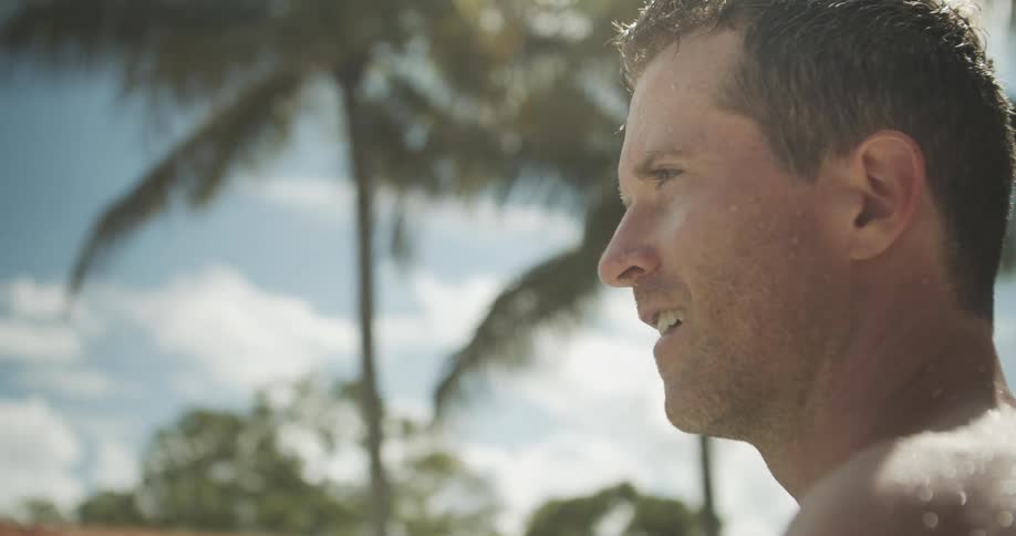 A close side view shows a wet shirtless man smiling in warm sunshine with palm trees and a bright blue sky visible in the soft focus background
