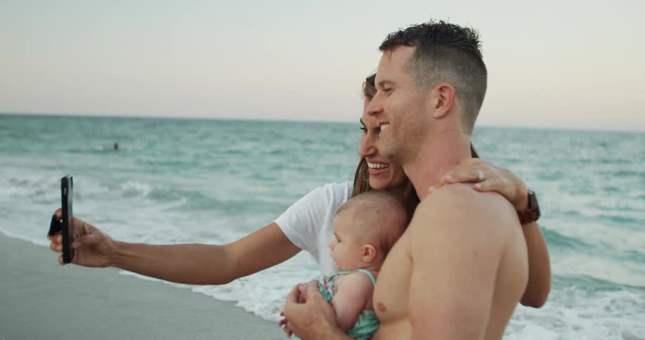 A shirtless man holding a baby stands with a woman wearing a white shirt on a sandy beach and the woman uses a phone to take a photo with the ocean and waves in the background