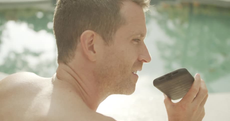 A shirtless man seen from over his shoulder and in profile speaks into his phone on speaker while sitting by a brightly sunlit pool