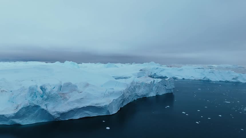 A sheer blue iceberg wall borders calm dark water beneath a flat gray sky