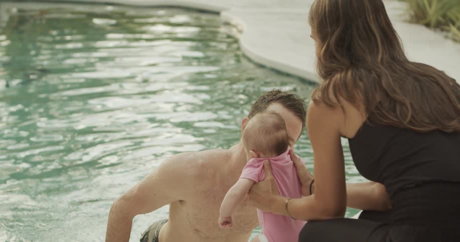 A woman in a black top holds a baby in a pink onesie while a man emerges from a clear swimming pool with a happy expression