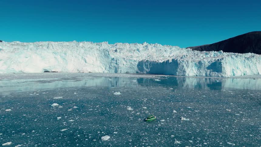 The video is an extreme wideangle shot of a massive glacier face that drops into a fjord creating a towering wall of white and blue ice in the foreground a small dark boat floats on the water filled with chunks of ice providing a sense of sc