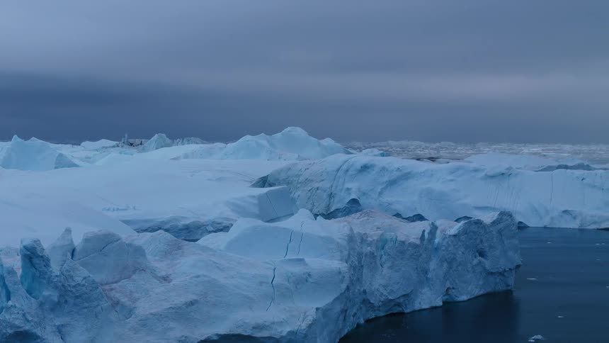 A huge field of white and blue icebergs and large ice masses floats on dark water under a gray cloudfilled sky moving slowly across the frame