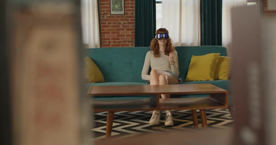 A young red haired woman wearing a light top shorts and a virtual reality headset sits on a teal sofa gesturing with her hand in a bright living room