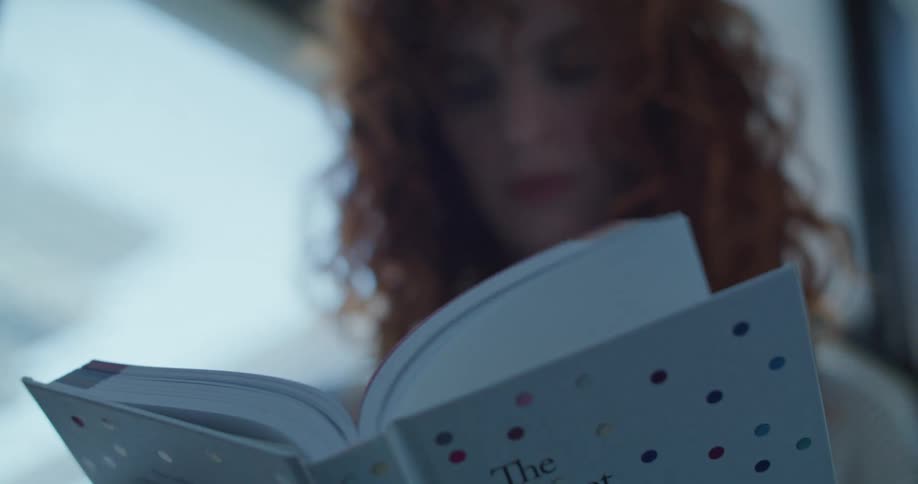 A close view of a woman with red curly hair reading a small polka dot patterned book with soft light coming from behind her