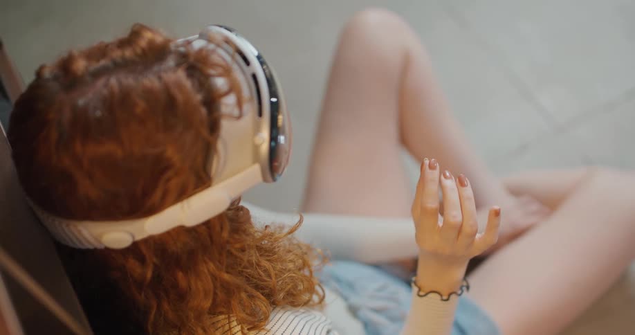 A redhaired woman wearing a vr set reclines on the floor with one hand raised appearing relaxed