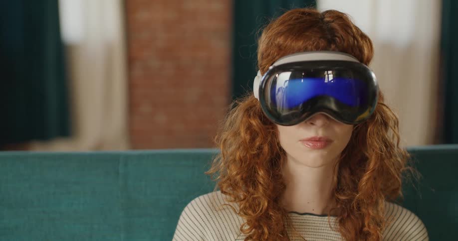 A close view shows a red haired woman wearing a light colored top and a sleek gray virtual reality headset with bright blue mirrored lenses