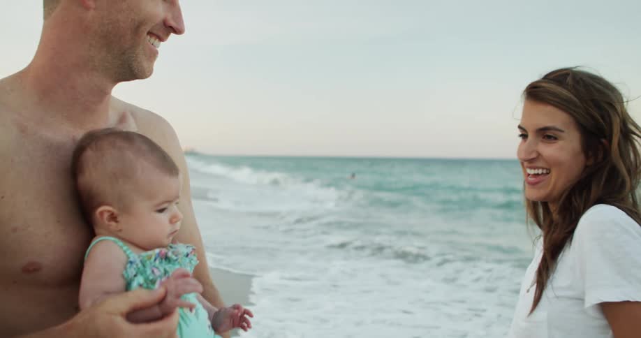 A man with a warm genuine smile holds a baby in a mint green swimsuit close to his chest on a sandy beach a womans profile is visible on the right also smiling as they all enjoy a bright day by the ocean