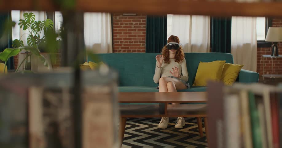 A young woman sits on a couch in her living room at midday wearing a vr headset and using hand gestures