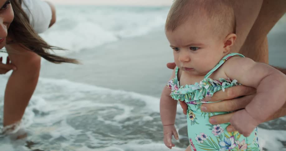 A close view of a baby in a floral swimsuit being held by a parent at the waters edge of a sandy beach with a woman in a white top smiling in the blurred background