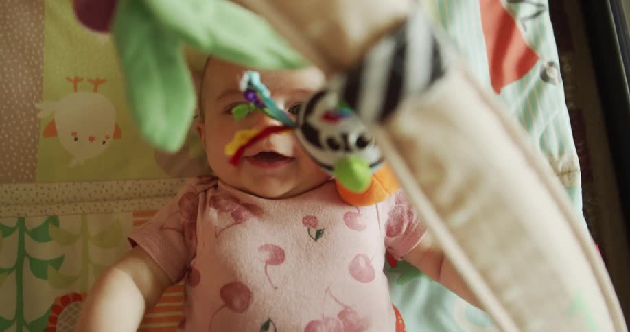 A baby in a pink onesie lies on a patterned play mat peeking past a dangling toy