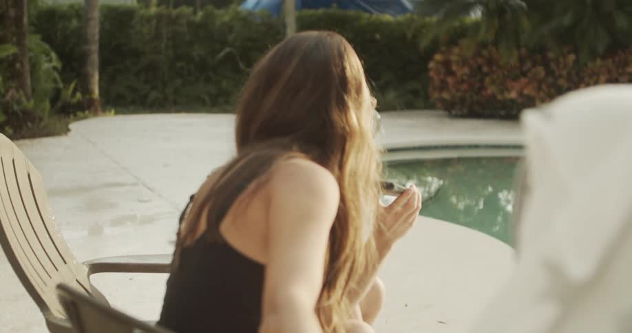 A medium closeup shot shows a man smiling and looking up from his smartphone while relaxing outdoors by a swimming pool he is sitting next to a woman with long hair who is smiling and facing the foreground