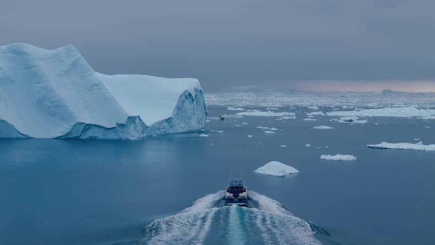 A high angle shot from behind shows a small blue and white motorboat cruising through dark arctic waters leaving a wake as it passes giant icebergs under a gray overcast sky