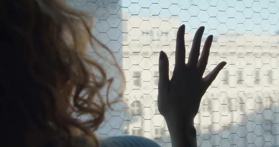 Woman with curly hair looks out a window while placing a hand on the mesh covering it