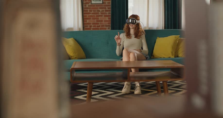 The video shows a woman with curly red hair sitting on a teal couch in a living room wearing a white vrmr headset she is seen gesturing with her right hand as if interacting with a virtual object or menu deeply engaged in the mixed reality env
