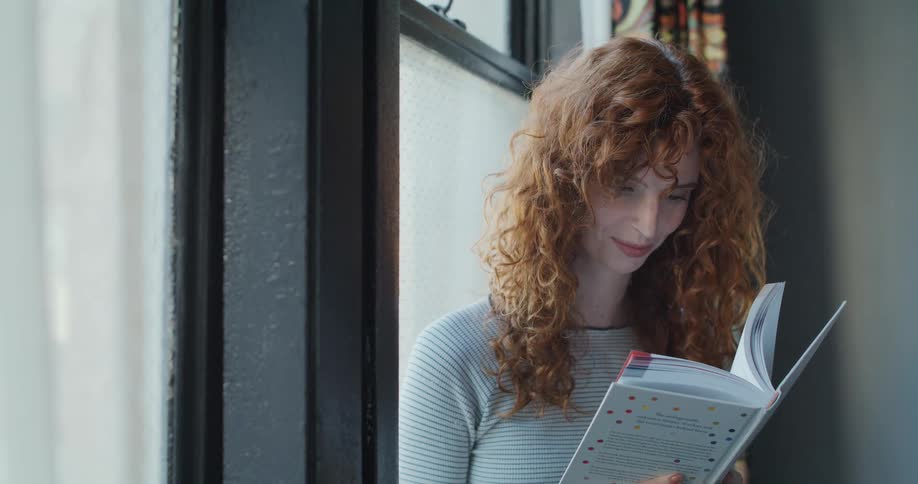 A smiling woman reads a book by a large indoor window