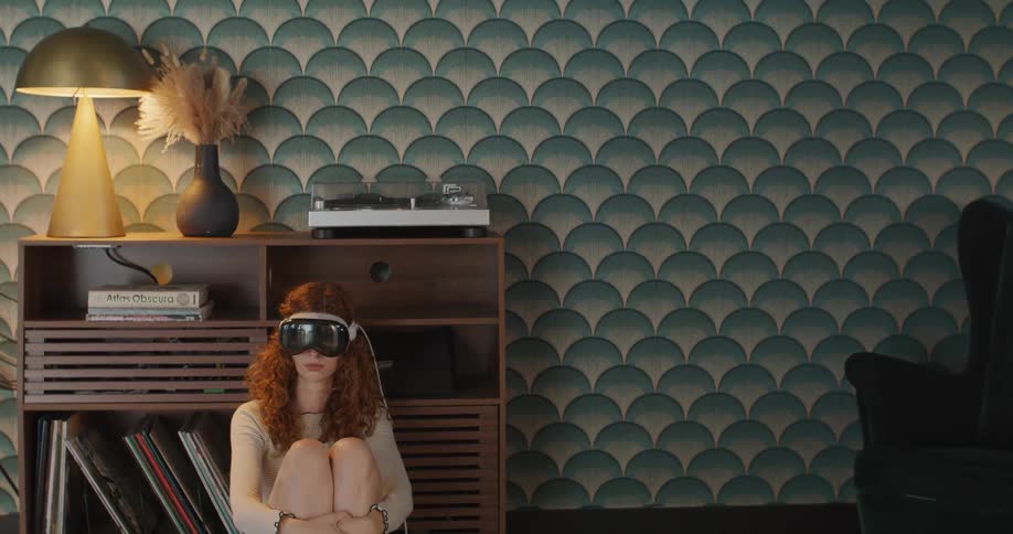The video shows a young woman with curly red hair sitting on a wooden storage unit hugging her knees to her chest while wearing a white virtual reality headset the wall behind her is covered in a striking tealandgreen scalloped patterned wa